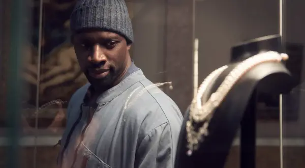 Omar Sy, protagonista di film e programmi TV, osserva con astuzia una collana di perle in una vetrina.