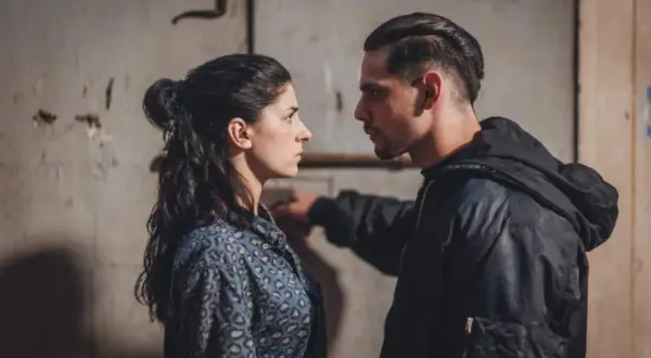 Una donna e un uomo si guardano intensamente. Lei indossa una camicia maculata, lui una giacca con cappuccio. Scena tesa, forse dalla serie "Rosa elettrica".