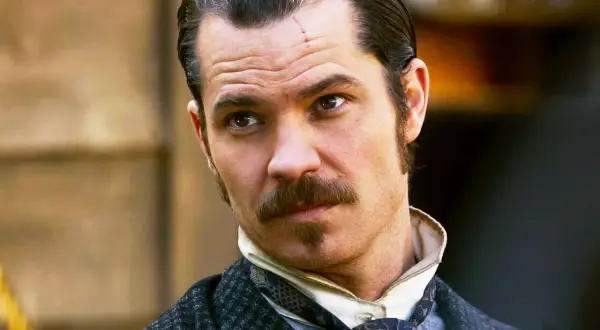 Timothy Olyphant, con cicatrice sulla fronte, baffi e basette, in abiti d'epoca, un volto iconico dei suoi film e programmi TV.
