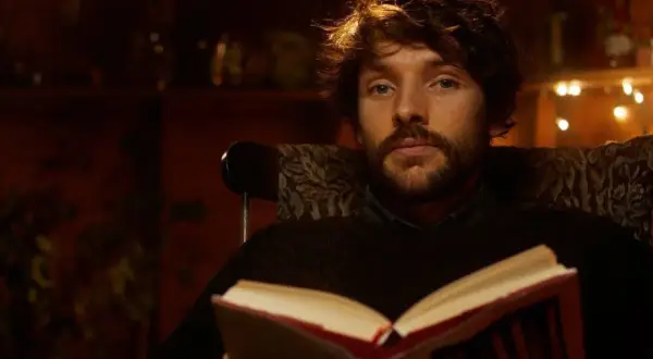 Un giovane uomo con barba e capelli castani, parte del cast di Merlin, legge un libro aperto seduto su una poltrona.