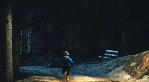 Bambino corre in un sentiero illuminato, in fondo al bosco, verso un misterioso oggetto luminoso nel cielo.