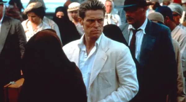 Willem Dafoe in un film, con un completo bianco, circondato da persone in abiti tradizionali e un uomo con cappello.