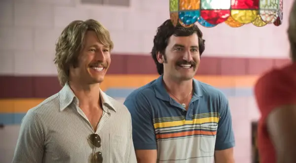 Due uomini sorridono, uno con capelli biondi e baffi, l'altro con capelli scuri e baffi. Scena da un film di Glen Powell.