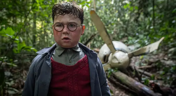 Ragazzo con occhiali e segni sul viso, in una foresta, vicino a un relitto d'aereo. Scena da "Il signore delle mosche serie tv".