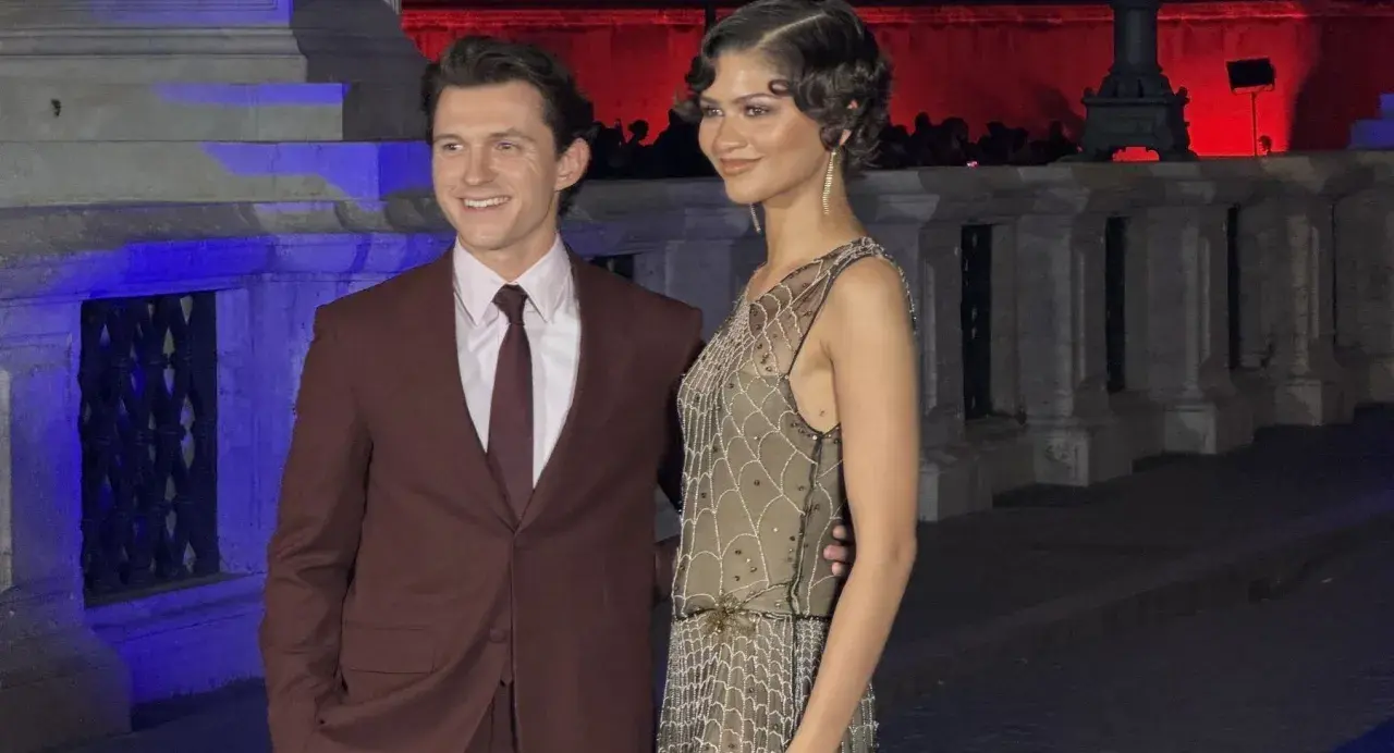 Tom Holland e Zendaya, una coppia iconica, sorridono alla telecamera. Lui indossa un completo bordeaux, lei un abito scintillante.