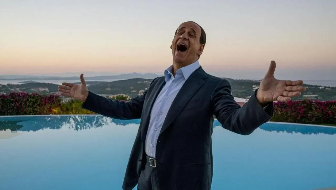Toni Servillo nei panni di Berlusconi, con le braccia aperte e la bocca spalancata, celebra un momento di gioia davanti a una piscina e al mare al tramonto.