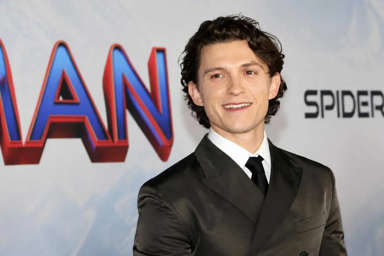 Tom Holland sorride, con indosso un abito scuro. Dietro di lui, il logo di Spider-Man. Chissà quanti anni ha Tom Holland?