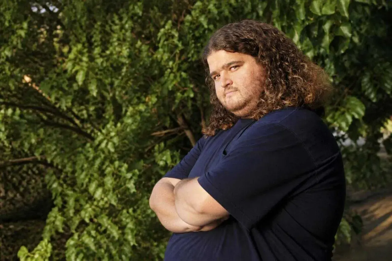 Jorge Garcia, noto per i suoi ruoli in film e programmi TV, con i suoi iconici capelli ricci e una maglietta blu, posa con le braccia conserte.