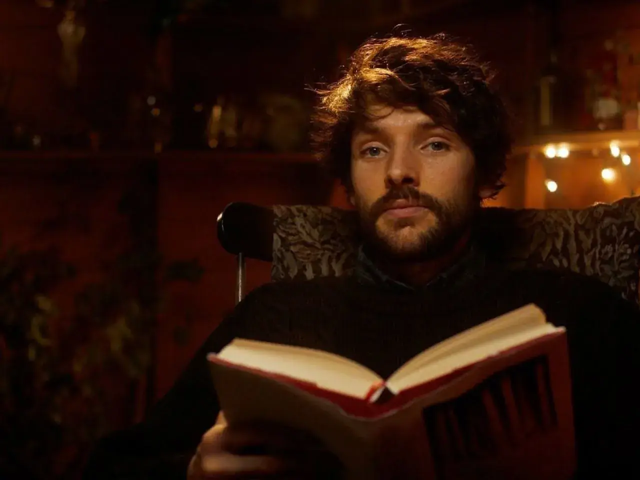 Un giovane uomo con barba e capelli castani, parte del cast di Merlin, legge un libro aperto seduto su una poltrona.