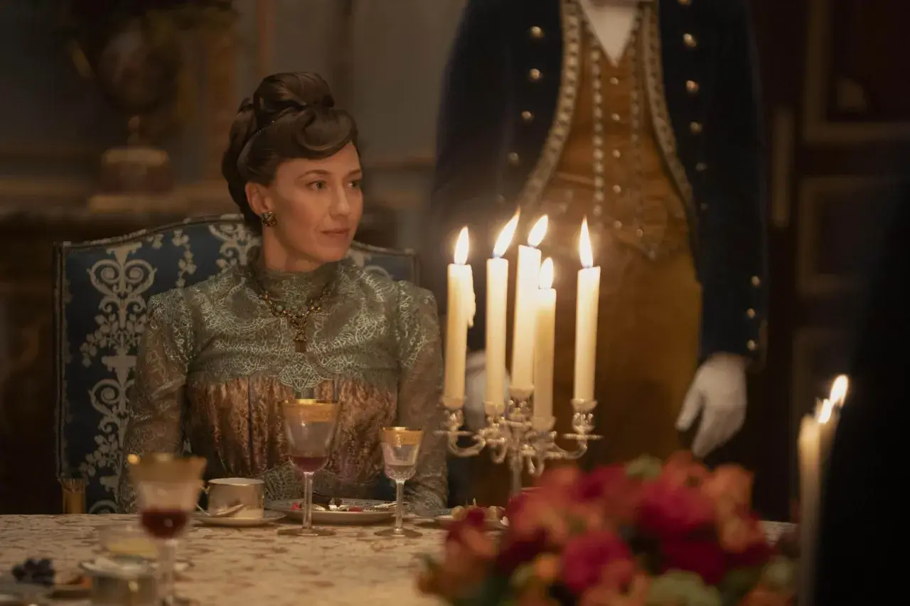 Una donna elegante, in abito d'epoca, siede a tavola durante un sontuoso banchetto, un'immagine che evoca lo sfarzo e le tensioni sociali della Gilded Age.