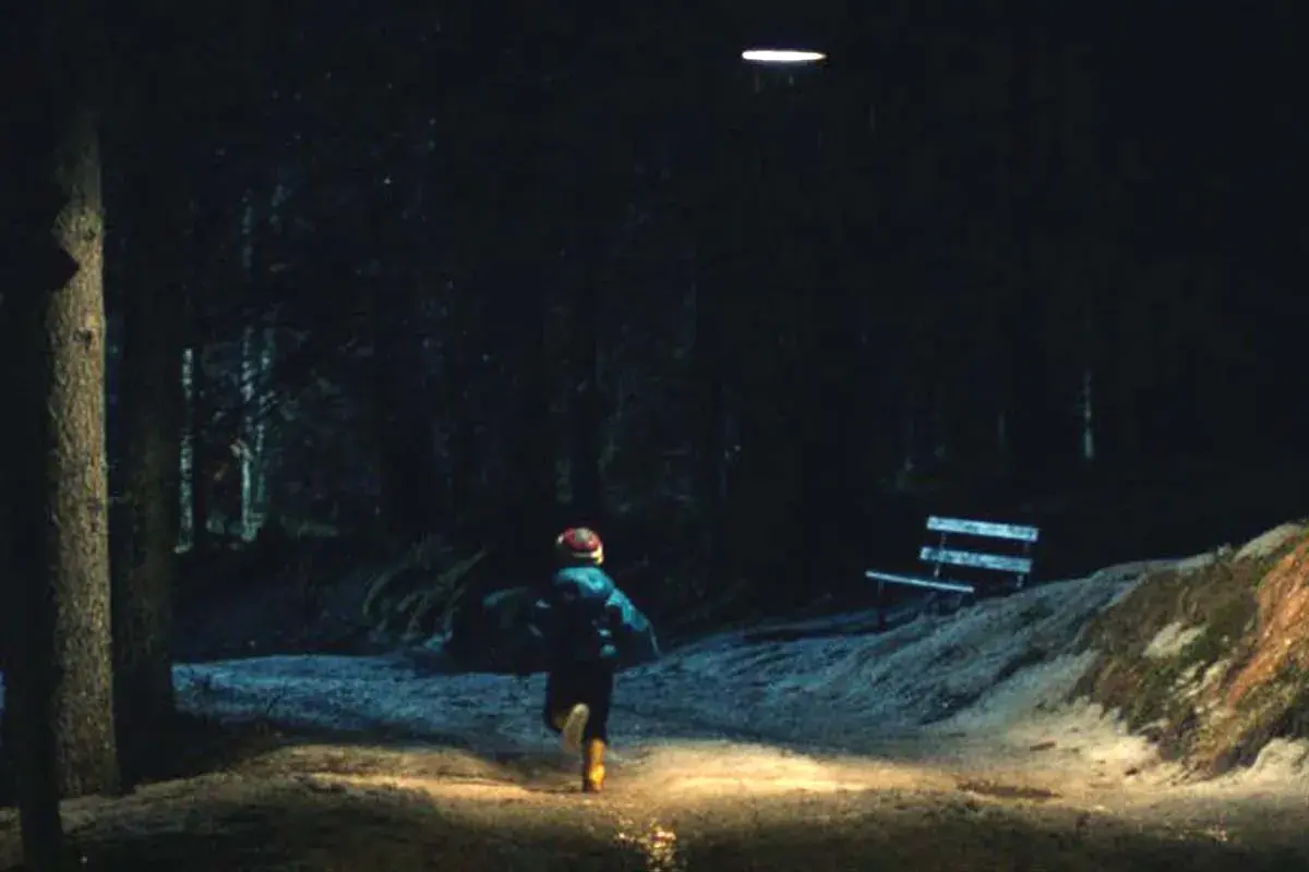Bambino corre in un sentiero illuminato, in fondo al bosco, verso un misterioso oggetto luminoso nel cielo.