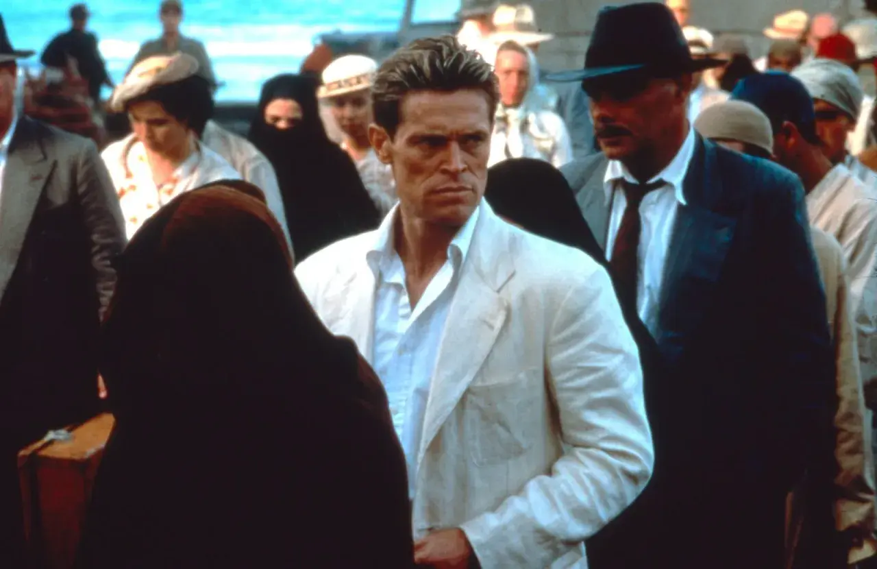 Willem Dafoe in un film, con un completo bianco, circondato da persone in abiti tradizionali e un uomo con cappello.