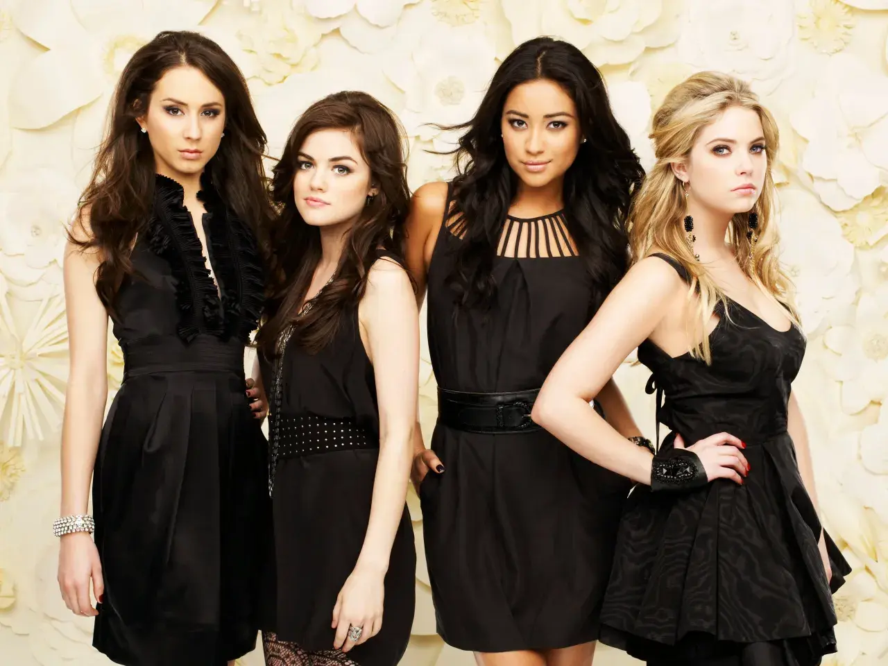 Il cast di Pretty Little Liars posa in abiti eleganti neri su uno sfondo floreale.