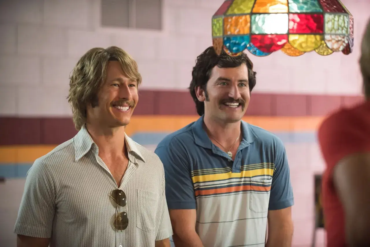 Due uomini sorridono, uno con capelli biondi e baffi, l'altro con capelli scuri e baffi. Scena da un film di Glen Powell.