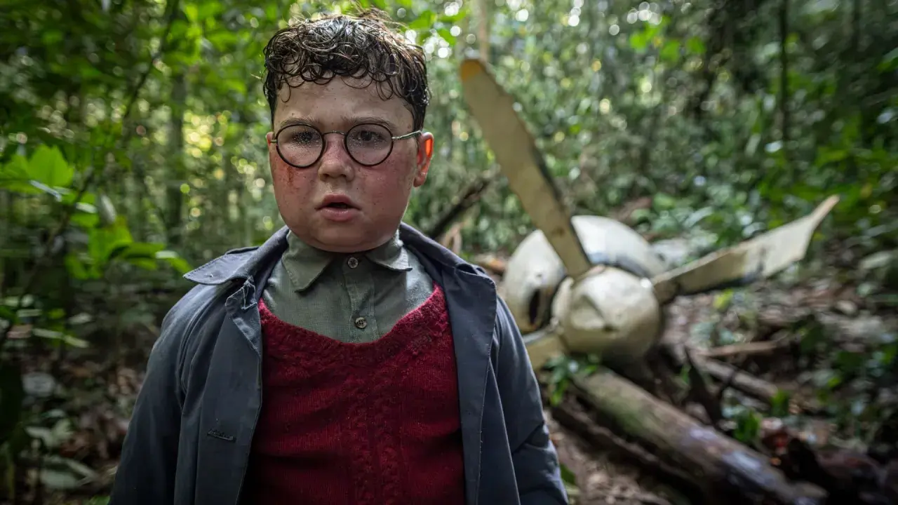 Ragazzo con occhiali e segni sul viso, in una foresta, vicino a un relitto d'aereo. Scena da "Il signore delle mosche serie tv".