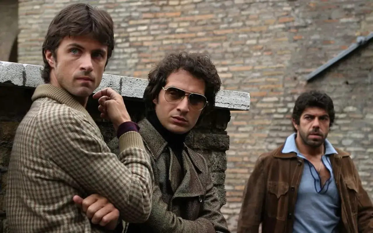 Il cast di Romanzo Criminale - La Serie: tre uomini con abiti anni '70 appoggiati a un muro di mattoni.