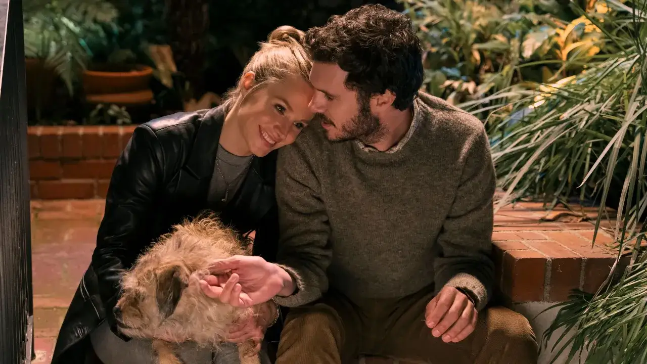 Coppia affettuosa con un cane, in un'ambientazione esterna. Adam Brody, noto per i suoi film e programmi TV, &egrave; in primo piano.