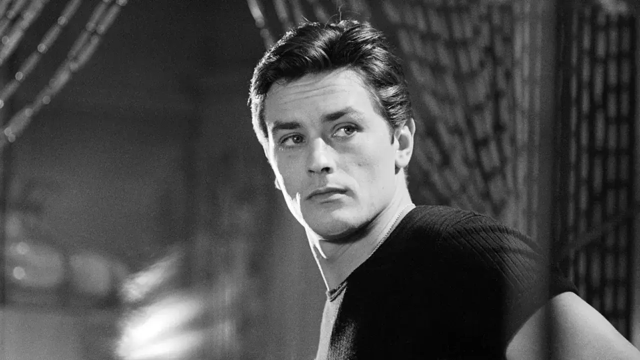 Alain Delon da ragazzo, con sguardo intenso e capelli scuri pettinati all'indietro, indossa una maglia a maniche corte.