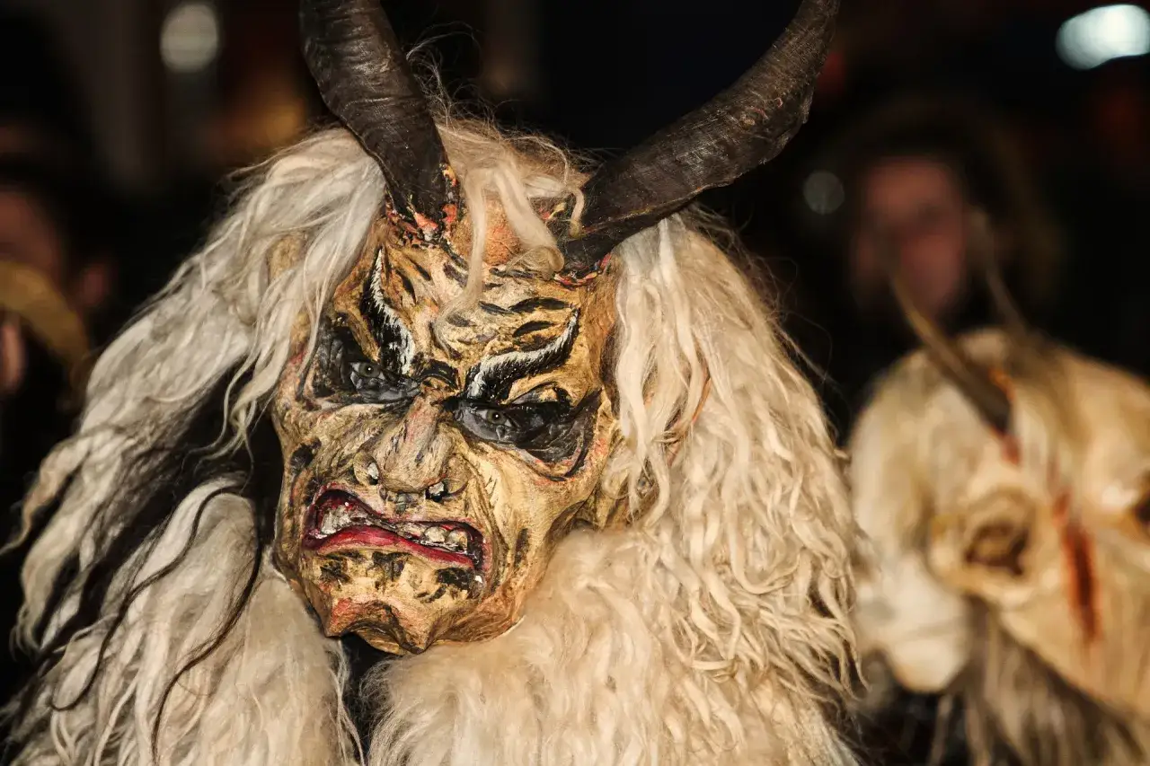 Maschera terrificante con corna e folta pelliccia bianca, sembra emergere in fondo al bosco, con altre figure sfocate sullo sfondo.