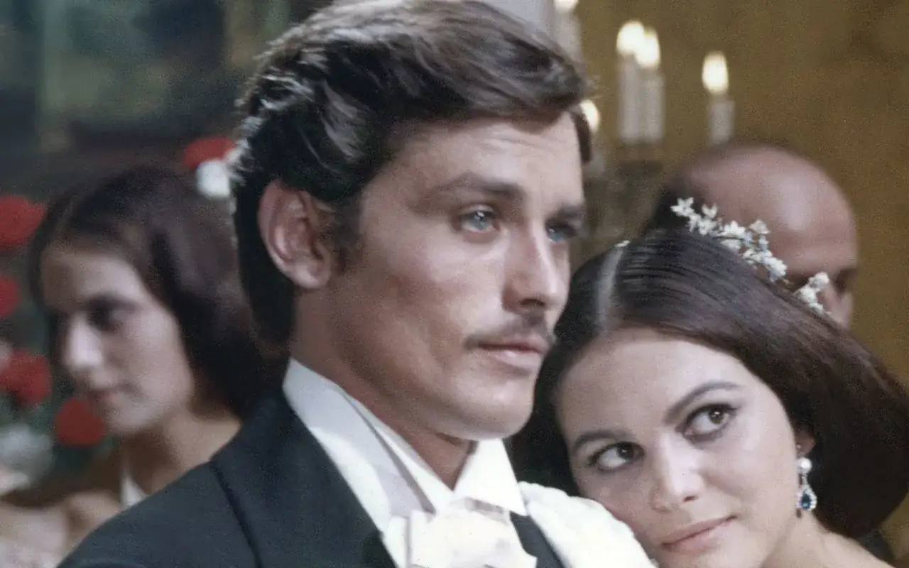 Alain Delon oggi, con un'espressione pensierosa, accanto a una donna con fiori tra i capelli.