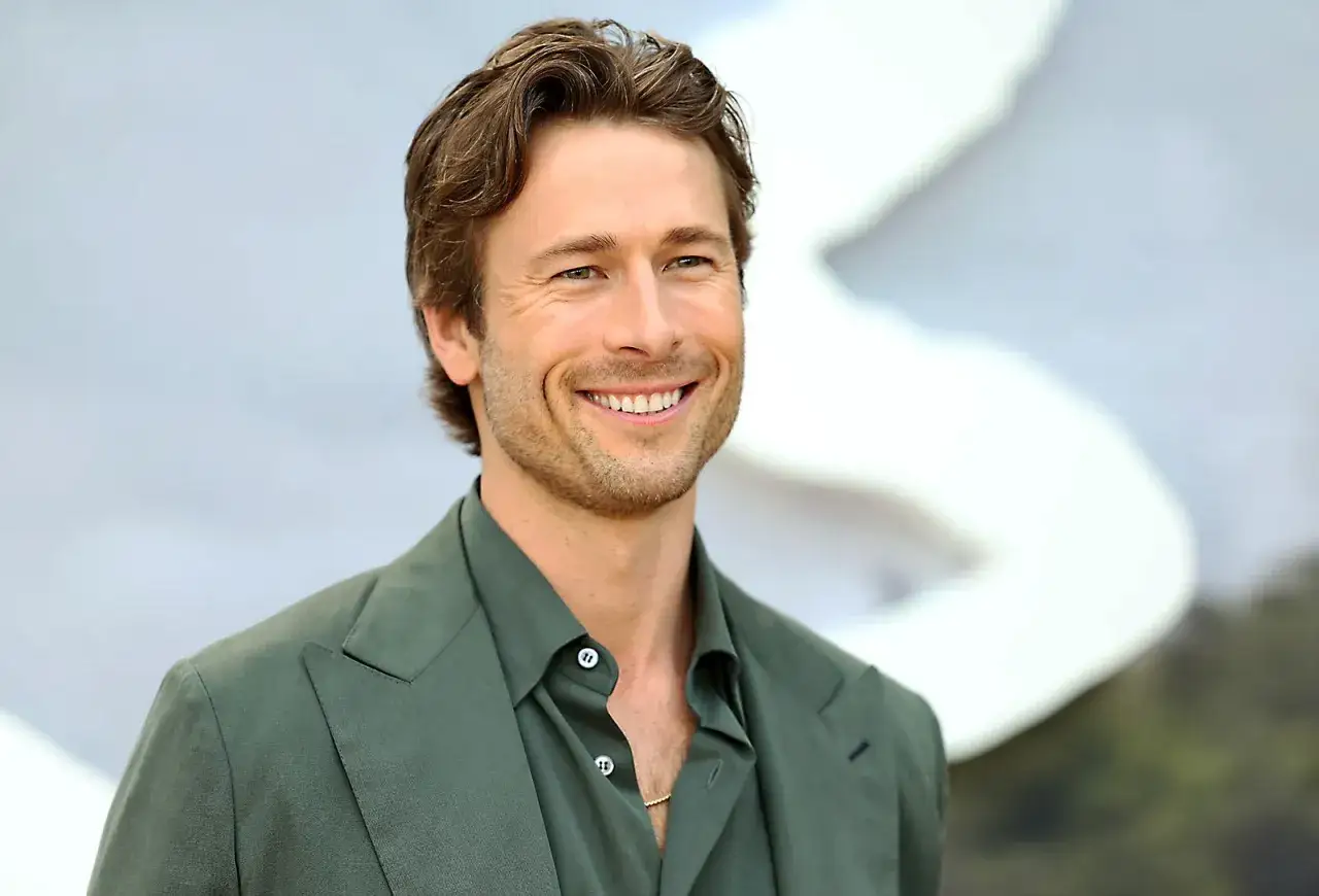 Glen Powell sorride con un completo verde, un look che ricorda i suoi film.