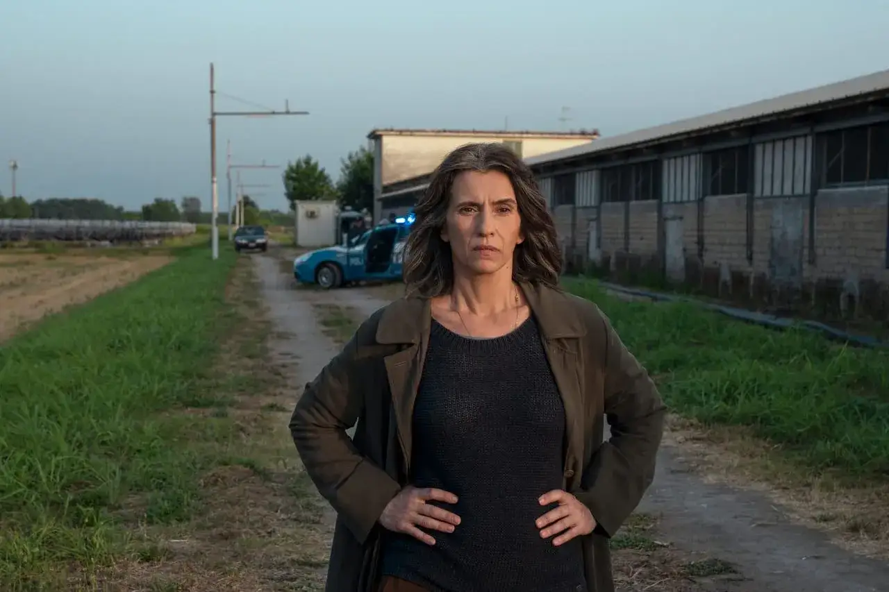 Donna con mani sui fianchi, in un'ambientazione rurale con auto della polizia. Scena tipica delle serie tv poliziesche.