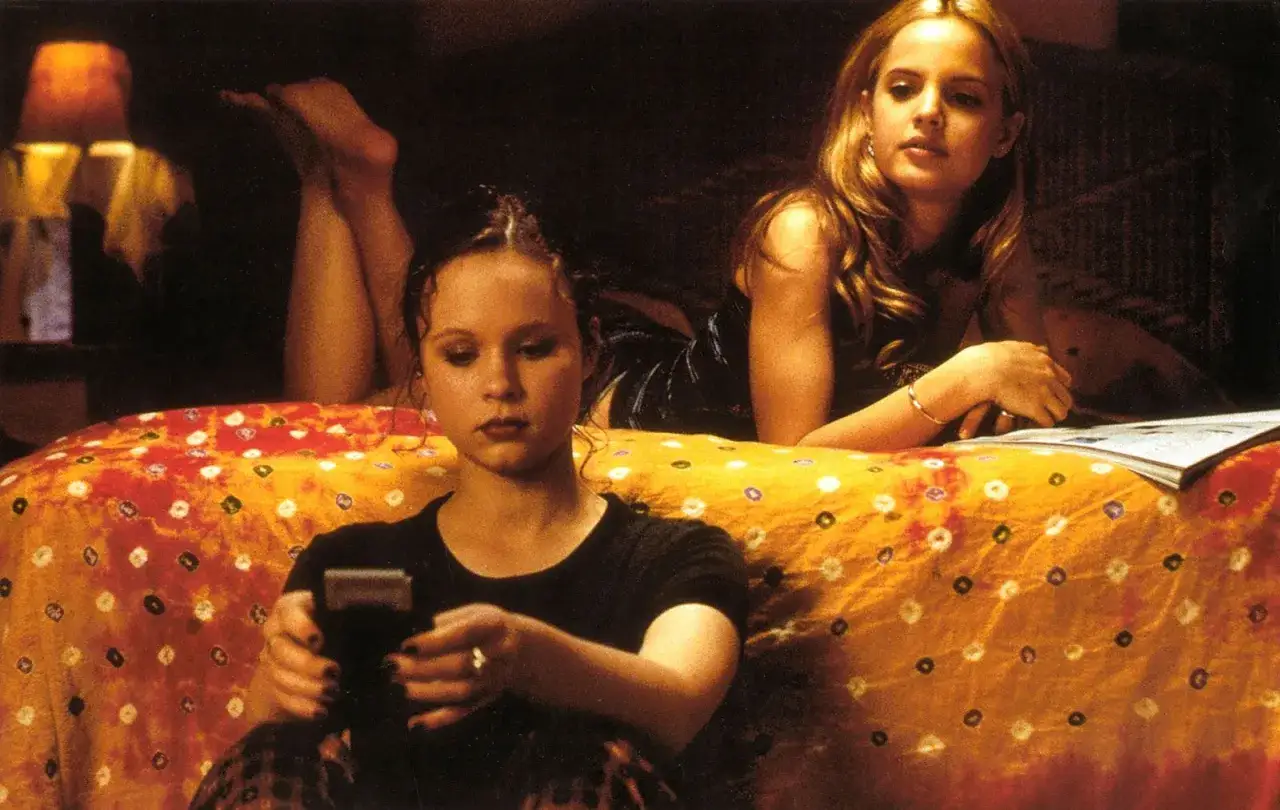 Il cast di American Beauty: due giovani donne su un letto colorato, una guarda un telefono, l'altra legge.