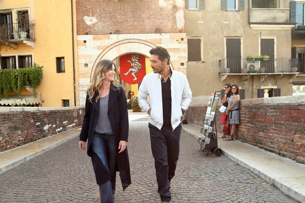 Coppia passeggia su un ponte acciottolato, mentre ero via, con edifici storici e un affresco sullo sfondo.