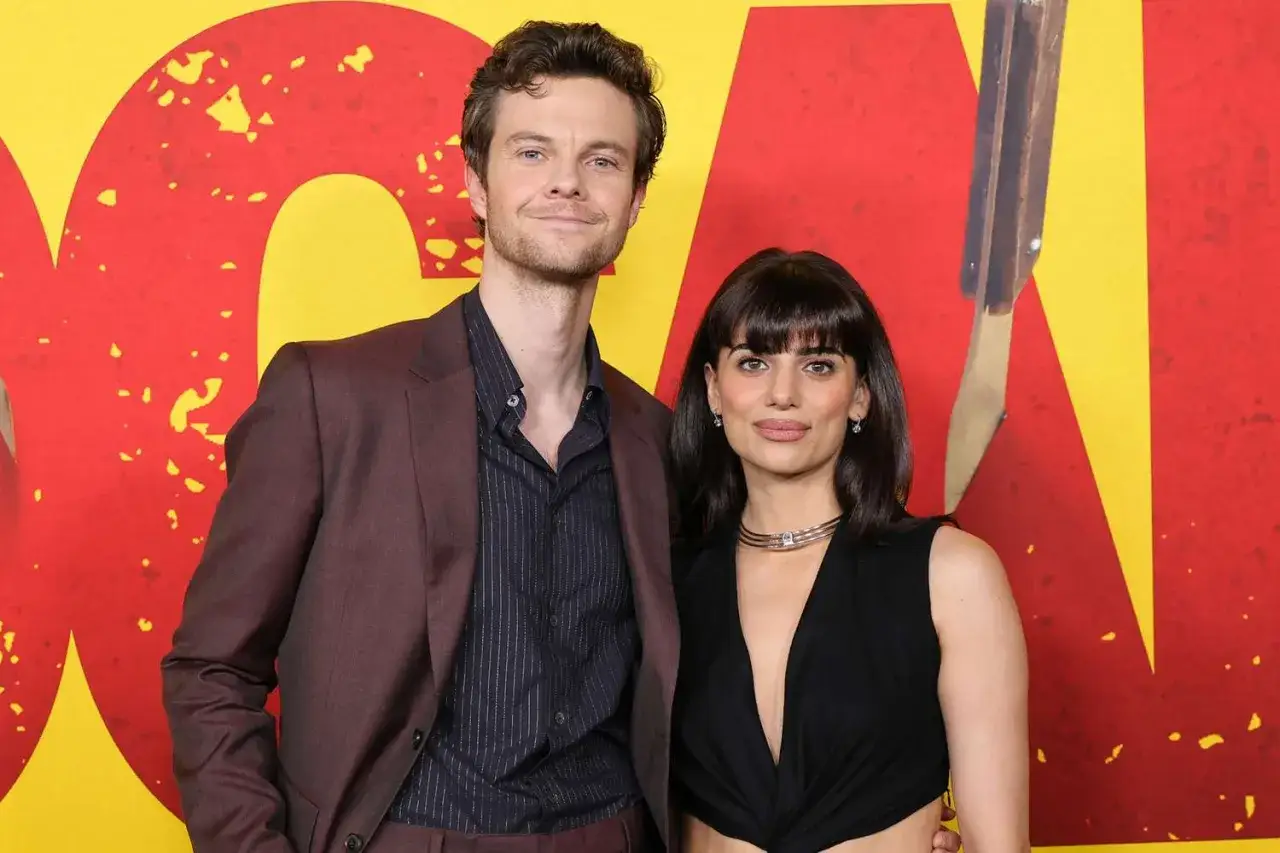 Jack Quaid con la sua fidanzata, sorridono davanti a uno sfondo rosso e giallo.