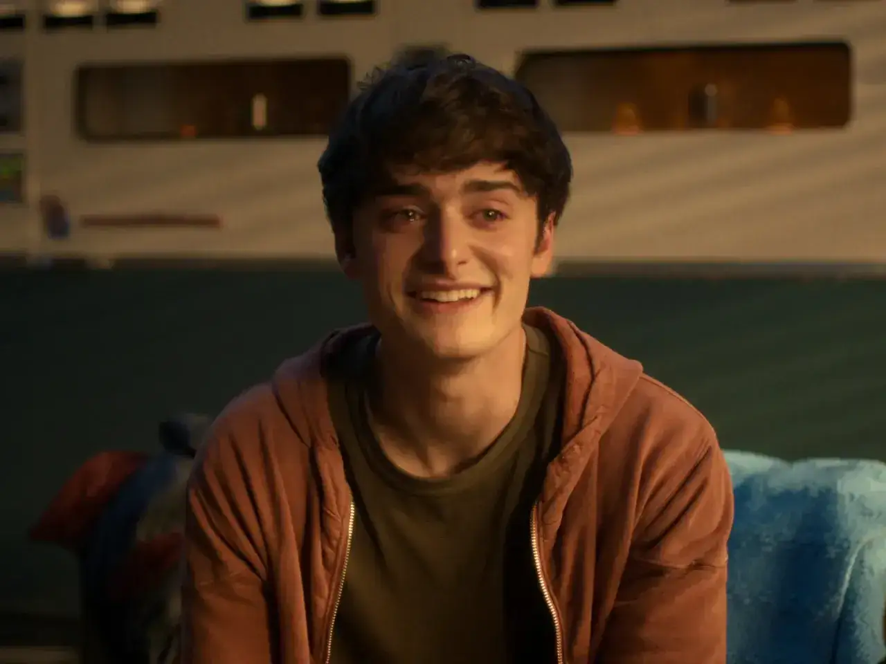 Noah Schnapp sorride, forse pensando al suo fidanzato. Indossa una felpa con cappuccio color ruggine.