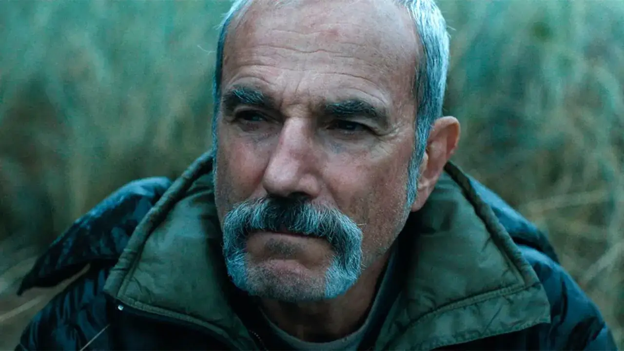 Primo piano di Daniel Day-Lewis in un film, con baffi grigi e sguardo intenso, immerso nella natura.