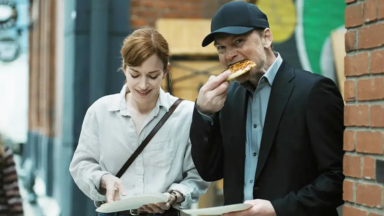 Un uomo con cappellino mangia una fetta di pizza, mentre una donna sorride con un piatto in mano. Scene dalla serie