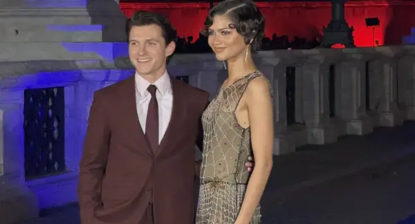 Tom Holland e Zendaya, una coppia iconica, sorridono alla telecamera. Lui indossa un completo bordeaux, lei un abito scintillante.
