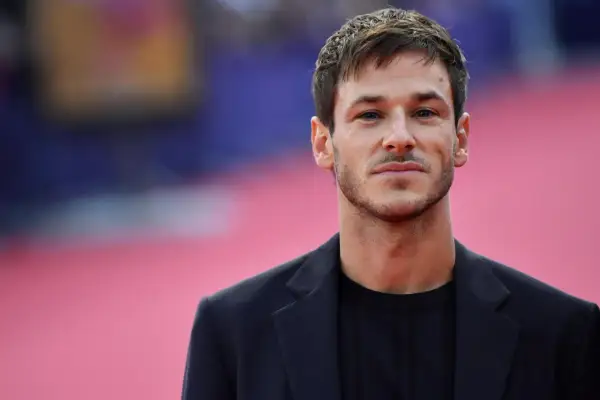 Ritratto di Gaspard Ulliel, attore morto oggi, con un blazer scuro su sfondo rosa sfocato.