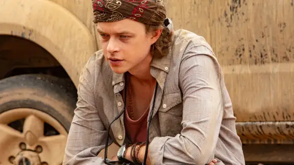 Dane DeHaan, con bandana e collana, pensieroso accanto a un veicolo fangoso in un film.