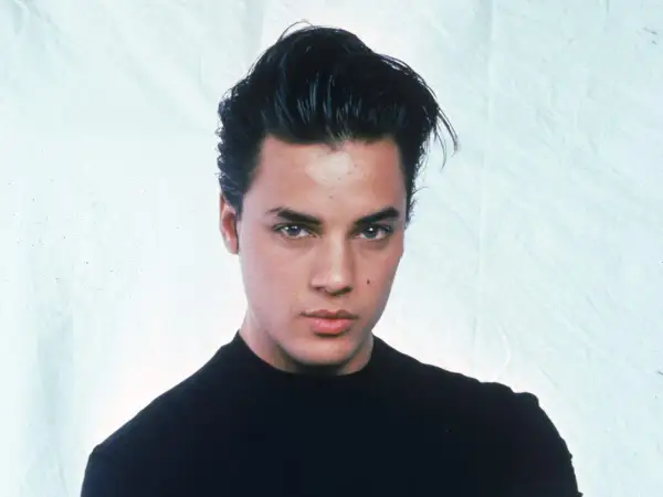 Ritratto di Nick Kamen, un giovane uomo con capelli scuri pettinati all'indietro e un maglione nero. La sua espressione è seria, mentre si riflette sulla sua **malattia**.