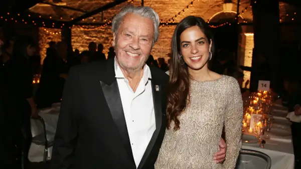 Alain Delon oggi, sorridente con una giovane donna elegante, durante un evento serale illuminato da lucine.
