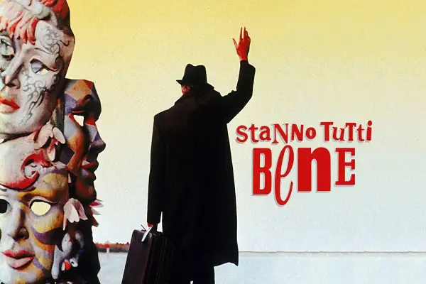 Un uomo con cappello e valigia saluta, circondato da maschere colorate. Il titolo del film "Stanno tutti bene" campeggia in rosso.