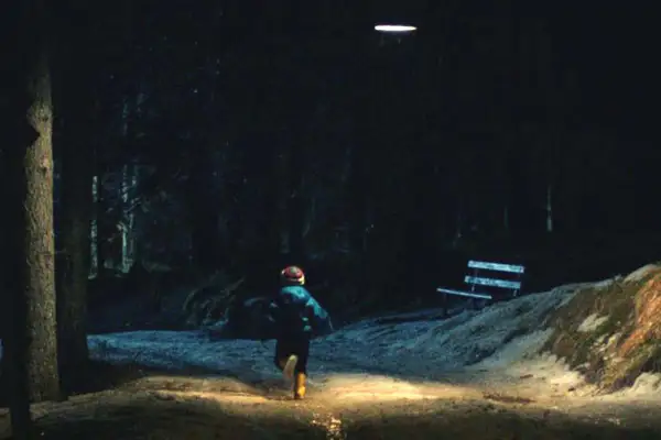 Bambino corre in un sentiero illuminato, in fondo al bosco, verso un misterioso oggetto luminoso nel cielo.