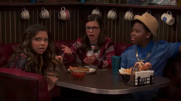 Benjamin Flores Jr. sorride con un cappello e una collana d'oro, mentre due ragazze discutono al tavolo di un diner.