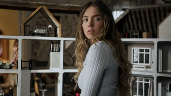 Una giovane donna con lunghi capelli castani guarda intensamente, circondata da case delle bambole. Sembra una di famiglia, forse la housemaid che osserva la vita degli altri.